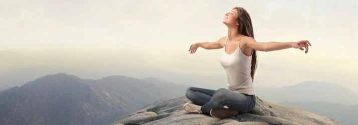 A person is seated on a rocky surface at a high elevation, with arms extended outward in an open posture. The background features a panoramic view of distant mountains under a soft, hazy sky, creating a serene and natural setting.